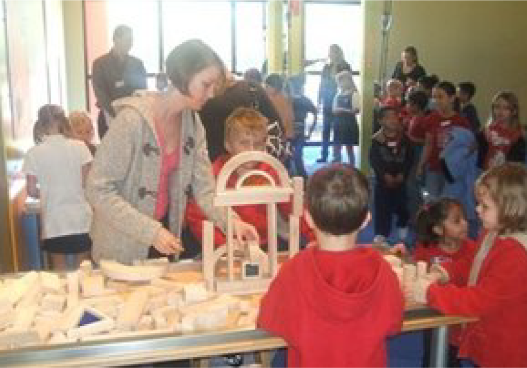 Asset 76@4x Children with teacher constructing items with blocks on table in museum.